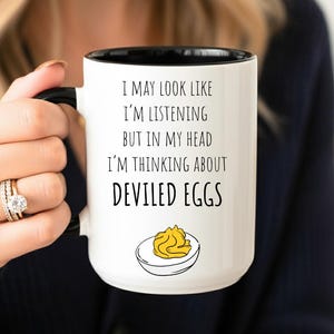 May include: White ceramic mug with a black interior and handle. The mug features the text "I MAY LOOK LIKE I'M LISTENING BUT IN MY HEAD I'M THINKING ABOUT DEVILED EGGS" with a graphic of a deviled egg.