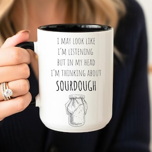 May include: White ceramic mug with a black interior and handle. The mug features the text "I MAY LOOK LIKE I'M LISTENING BUT IN MY HEAD I'M THINKING ABOUT SOURDOUGH" and a drawing of a sourdough starter jar.
