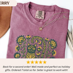 May include: Berry-colored t-shirt with a cat and floral graphic. The design includes the text "chinga la". A gold necklace with a pendant rests on the shirt. The shirt has a Comfort Colors tag.