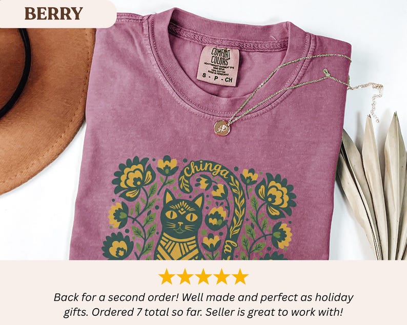 May include: Berry-colored t-shirt with a cat and floral graphic. The design includes the text "chinga la". A gold necklace with a pendant rests on the shirt. The shirt has a Comfort Colors tag.