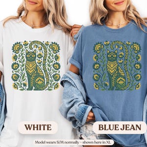 May include: White and blue jean t-shirts featuring a green cat design with floral accents. The cat has a decorative pattern and the text "chinga la migra". The shirts are a relaxed fit.