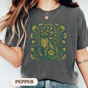 May include: Dark gray t-shirt with a green and gold cat design surrounded by floral elements. The text "chinga la migra" is arched above and below the cat illustration. The shirt is a relaxed fit.