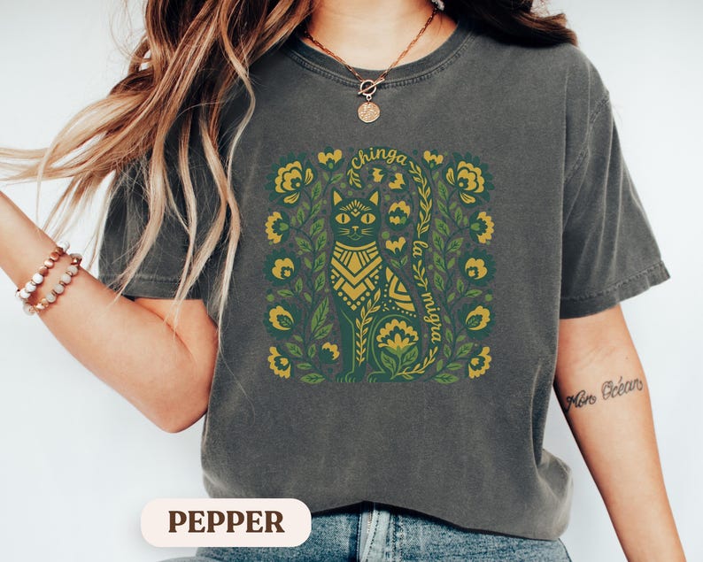 May include: Dark gray t-shirt with a green and gold cat design surrounded by floral elements. The text "chinga la migra" is arched above and below the cat illustration. The shirt is a relaxed fit.