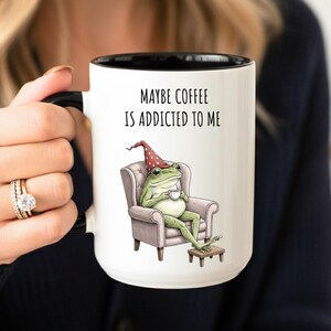 May include: White ceramic coffee mug with a black interior and handle. The mug features a whimsical illustration of a frog in a chair, wearing a hat and holding a cup. Text on the mug reads, "MAYBE COFFEE IS ADDICTED TO ME."