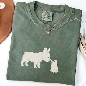 May include: A moss green short-sleeve t-shirt featuring a graphic of a cream-colored French Bulldog touching noses with a small white ghost. The shirt has a crew neckline and a Comfort Colors brand label printed on the inside collar.