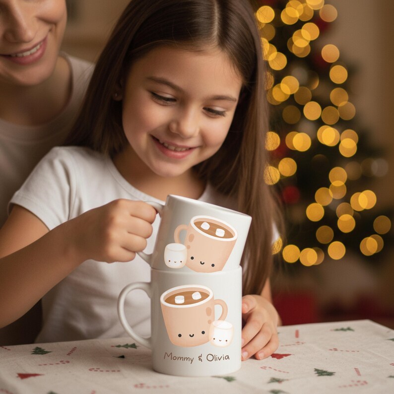 May include: Two white ceramic mugs stacked, each with a cartoon hot chocolate design and marshmallows. The bottom mug has the text "Mommy & Olivia". A young girl holds the mugs. The background includes a Christmas tree with bokeh lights.