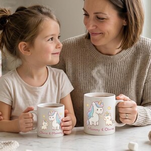 May include: Two white ceramic mugs featuring a unicorn design. One mug is held by a child and the other by an adult. The mugs have the text "Mommy & Olivia" and "Olivia & Mommy". The unicorns have pastel manes and tails.