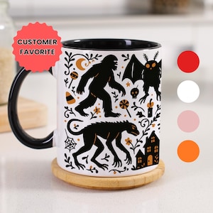 May include: White ceramic mug with a black handle and rim, featuring black silhouettes of cryptids and Halloween-themed illustrations. The mug includes the text "CUSTOMER FAVORITE" on a red starburst.