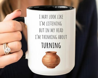 May include: White ceramic mug with a black interior and handle. The mug features the text "I MAY LOOK LIKE I'M LISTENING BUT IN MY HEAD I'M THINKING ABOUT TURNING" and a brown pottery illustration. The mug is held by a person.
