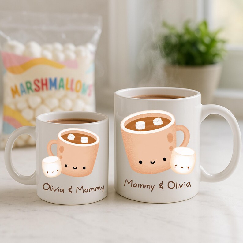 May include: Two white ceramic mugs with a hot chocolate design. Each mug features a cartoon illustration of a mug of hot chocolate with marshmallows and a smiling marshmallow. One mug says "Olivia & Mommy", the other says "Mommy & Olivia".