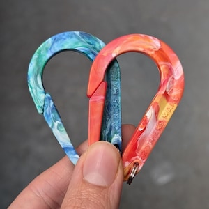 May include: Two carabiner clips are shown. One is blue and green, the other is red, orange, and yellow. The text "BROTHERS MAKE" is printed on the side of each clip. The clips are made of a marbled material.