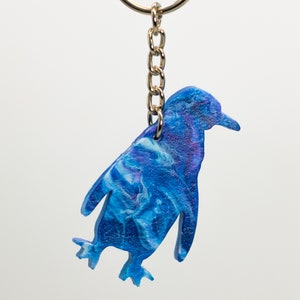 May include: A blue and white penguin-shaped keychain with a gold chain and ring. The penguin is painted with a swirling blue and white pattern.