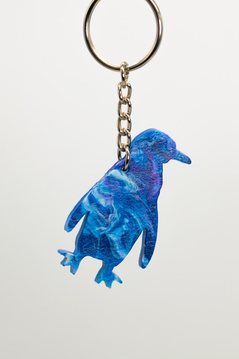 May include: A blue and white penguin-shaped keychain with a gold chain and ring. The penguin is painted with a swirling blue and white pattern.