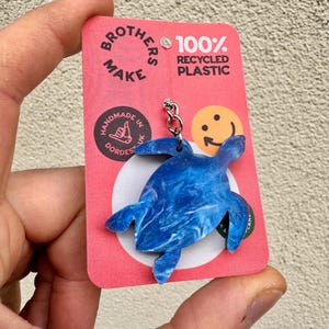 May include: Blue and white marbled plastic turtle keychain with a silver chain. The keychain is attached to a pink card with the text "Brothers Make 100% Recycled Plastic" and a smiley face.