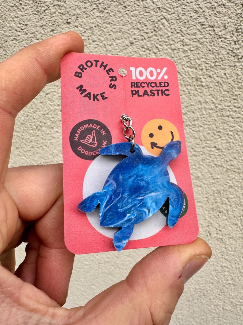 May include: Blue and white marbled plastic turtle keychain with a silver chain. The keychain is attached to a pink card with the text "Brothers Make 100% Recycled Plastic" and a smiley face.