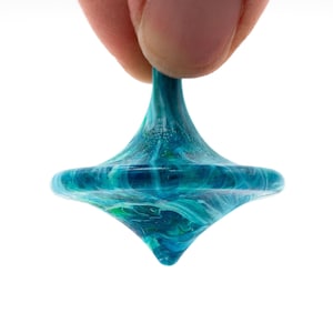 May include: A handcrafted, colorful spinning top with a swirling blue and green pattern. The top is held by a person's fingers against a white background. The toy has a rounded base and a pointed tip.