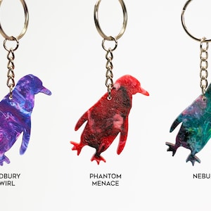 May include: Three penguin-shaped keychains with different colorful marbled patterns. The keychains are labeled "Cadbury Swirl", "Phantom Menace", and "Nebula".