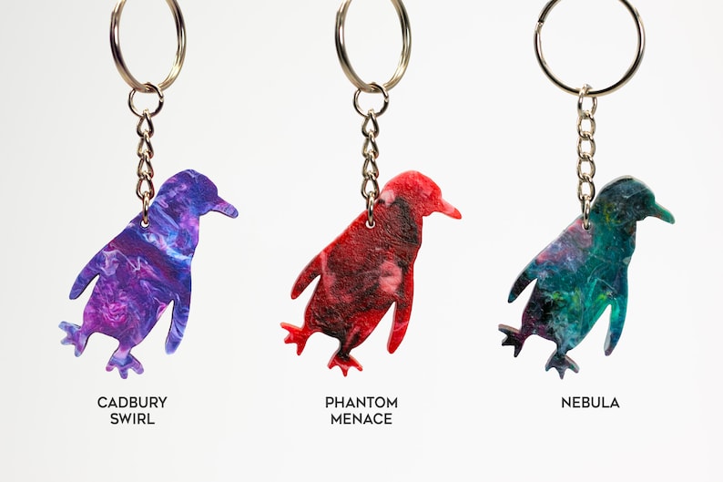 May include: Three penguin-shaped keychains with different colorful marbled patterns. The keychains are labeled "Cadbury Swirl", "Phantom Menace", and "Nebula".