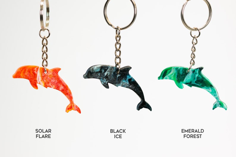 May include: Three dolphin keychains with different color patterns: orange and white, black and blue, and green and white. The keychains are labeled "Solar Flare", "Black Ice", and "Emerald Forest".