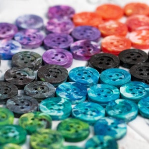 May include: A collection of colourful buttons in various shades of blue, purple, black, and green. The buttons are made of a marbled plastic material and have a unique, speckled appearance.