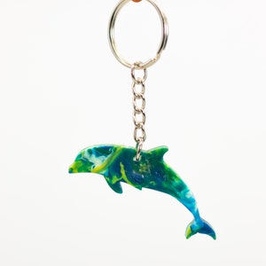 May include: A green, blue, and yellow dolphin keychain. The dolphin is jumping out of the water. The keychain is attached to a silver ring and chain.