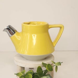 May include: A bright yellow ceramic teapot with a silver spout. The teapot has a white base and a handle. The teapot is sitting on a small, round, white wooden stand. Greenery is placed in front of the teapot.