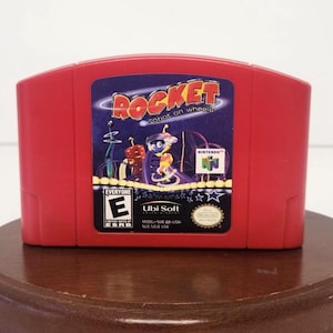 May include: A red Nintendo 64 game cartridge for "Rocket: Robot on Wheels." The label features a cartoon robot character and the game's title. The cartridge is marked "Everyone" by ESRB and "Ubi Soft."