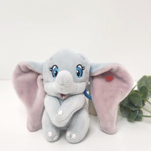 May include: A plush toy elephant in a seated position, featuring large, light pink ears and blue eyes with long eyelashes. The elephant is primarily gray with a small red heart on one ear. A blue loop is attached to the toy.
