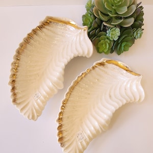 May include: Two white ceramic leaf-shaped dishes with gold trim. The dishes are slightly curved and have a textured surface. Each dish has a small white label with the text "81/2" and "564" on it.