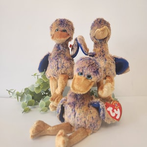 May include: Three purple and brown plush toy birds with blue wings. The birds have yellow beaks and black eyes. One bird has a Ty tag attached to its leg.