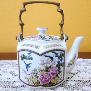 May include: White ceramic teapot with a floral and bird design. The teapot features a gold-colored handle and spout. The design includes pink peonies, blue birds, and other colorful floral elements.
