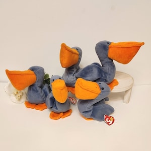 May include: Four gray and orange plush pelican beanie babies. The beanie babies are sitting on a small wooden table. The beanie babies have a Ty tag attached to one of them.