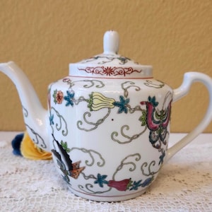 May include: A white porcelain teapot featuring a colorful pattern of butterflies, flowers, and swirling vines. The teapot has a matching lid with a small knob, a curved spout, and a handle. The design includes shades of red, yellow, and blue.