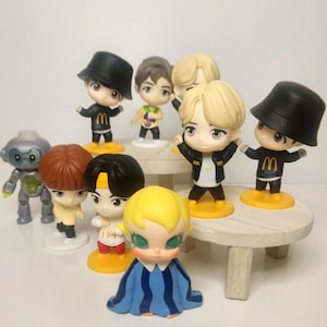 May include: A collection of small figurines, including several with black bucket hats and the letter "M" on their shirts. Other figures include a robot and a doll with blue and white striped clothing. The figurines are on a small, round wooden table.