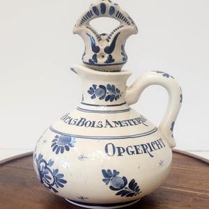 May include: A cream-colored ceramic decanter with a matching stopper, featuring blue floral patterns and the text Lucas Bols Amsterdam and Opgericht. The vessel has a rounded body, a curved handle, and a spout, decorated with small star motifs.