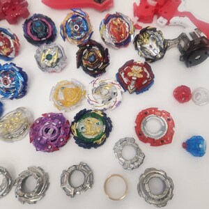 Puede incluir: Una colecci&oacute;n de coloridos trompos y lanzadores. Los trompos presentan varios dise&ntilde;os con detalles en rojo, azul, dorado y morado. Tambi&eacute;n hay varios lanzadores rojos y blancos, junto con un anillo dorado.