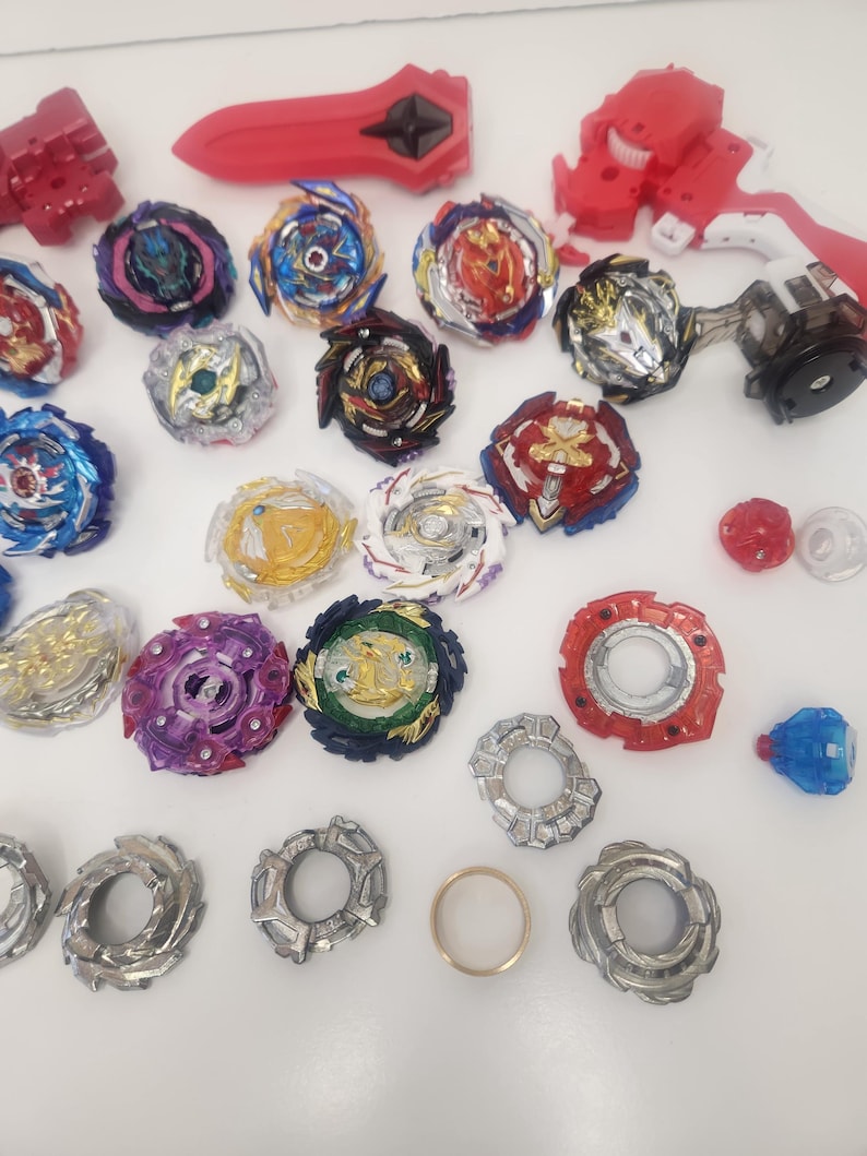 Puede incluir: Una colecci&oacute;n de coloridos trompos y lanzadores. Los trompos presentan varios dise&ntilde;os con detalles en rojo, azul, dorado y morado. Tambi&eacute;n hay varios lanzadores rojos y blancos, junto con un anillo dorado.