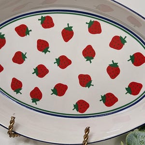 May include: Oval serving platter with a white background and a pattern of red strawberries with green stems. The platter has a blue and green border and gold-colored metal handles.