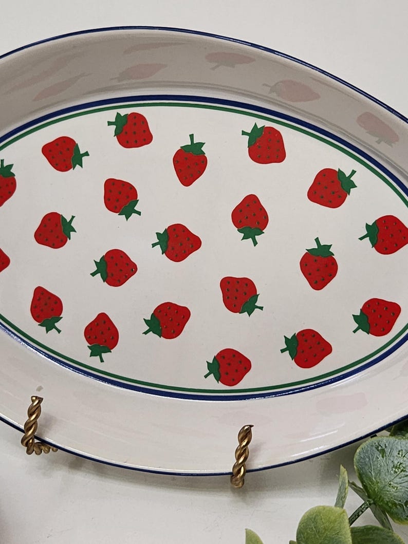 May include: Oval serving platter with a white background and a pattern of red strawberries with green stems. The platter has a blue and green border and gold-colored metal handles.