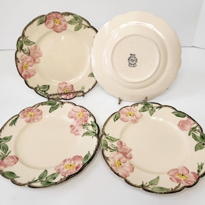 May include: A set of four cream-colored ceramic plates with scalloped edges, featuring hand-painted pink wild rose blossoms and green leaves. One plate is flipped over to show the back stamp reading Hand Painted Francisco USA.