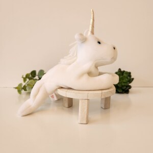 K&ouml;nnte beinhalten: Wei&szlig;es Pl&uuml;sch-Einhorn-Spielzeug mit goldenem Horn, das auf einem kleinen Holzhocker sitzt. Das Einhorn hat blaue Augen und eine wei&szlig;e M&auml;hne.