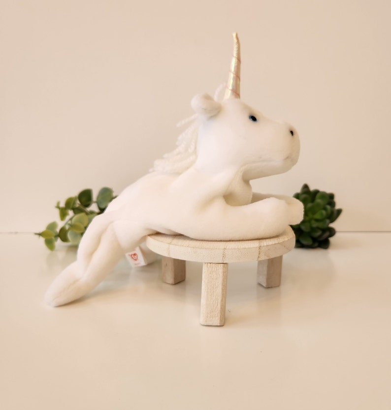 K&ouml;nnte beinhalten: Wei&szlig;es Pl&uuml;sch-Einhorn-Spielzeug mit goldenem Horn, das auf einem kleinen Holzhocker sitzt. Das Einhorn hat blaue Augen und eine wei&szlig;e M&auml;hne.