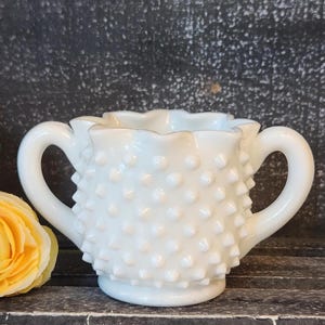 May include: A white hobnail creamer with two curved handles. The creamer has a ruffled rim and is covered in small, raised bumps. The creamer is a vintage piece, and is likely made of milk glass.