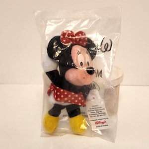 May include: A Minnie Mouse plush toy in a clear plastic bag. The toy has a red and white polka dot bow, a red and white polka dot skirt, and yellow shoes. The packaging includes the Kellogg's logo and "MADE IN CHINA".