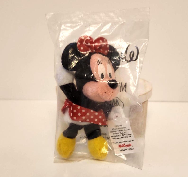 May include: A Minnie Mouse plush toy in a clear plastic bag. The toy has a red and white polka dot bow, a red and white polka dot skirt, and yellow shoes. The packaging includes the Kellogg's logo and "MADE IN CHINA".