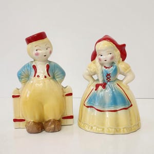May include: Two ceramic figurines depicting a pair of children. The left figure wears a red cap, blue jacket, and yellow trousers standing by a fence. The right figure wears a red hood, blue apron, and yellow dress with red lace-up details.