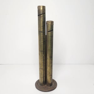 May include: A vintage brass coin sorter with two cylindrical tubes of different heights. The tubes have black markings indicating dollar amounts. The sorter is mounted on a circular base.