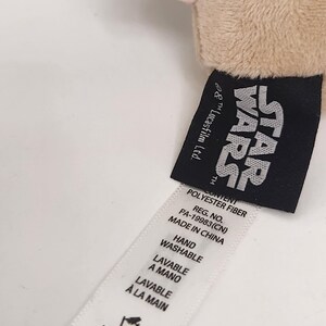 May include: A close-up of a plush toy with a Star Wars tag. The tag is black with white text and includes the Star Wars logo. A white tag with washing instructions is also visible.