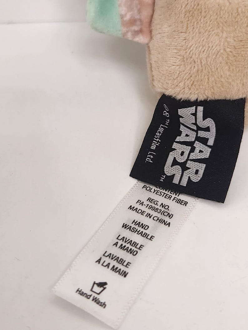 May include: A close-up of a plush toy with a Star Wars tag. The tag is black with white text and includes the Star Wars logo. A white tag with washing instructions is also visible.