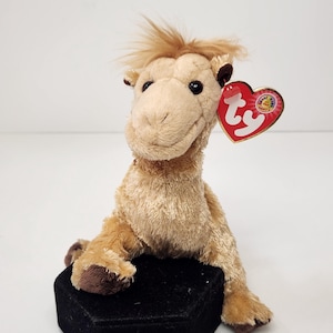 May include: A tan plush camel stuffed animal with a tuft of hair on its head and dark brown hooves. A red heart-shaped Ty tag is attached to the camel's left ear, featuring the white Ty logo and a small circular emblem with text.